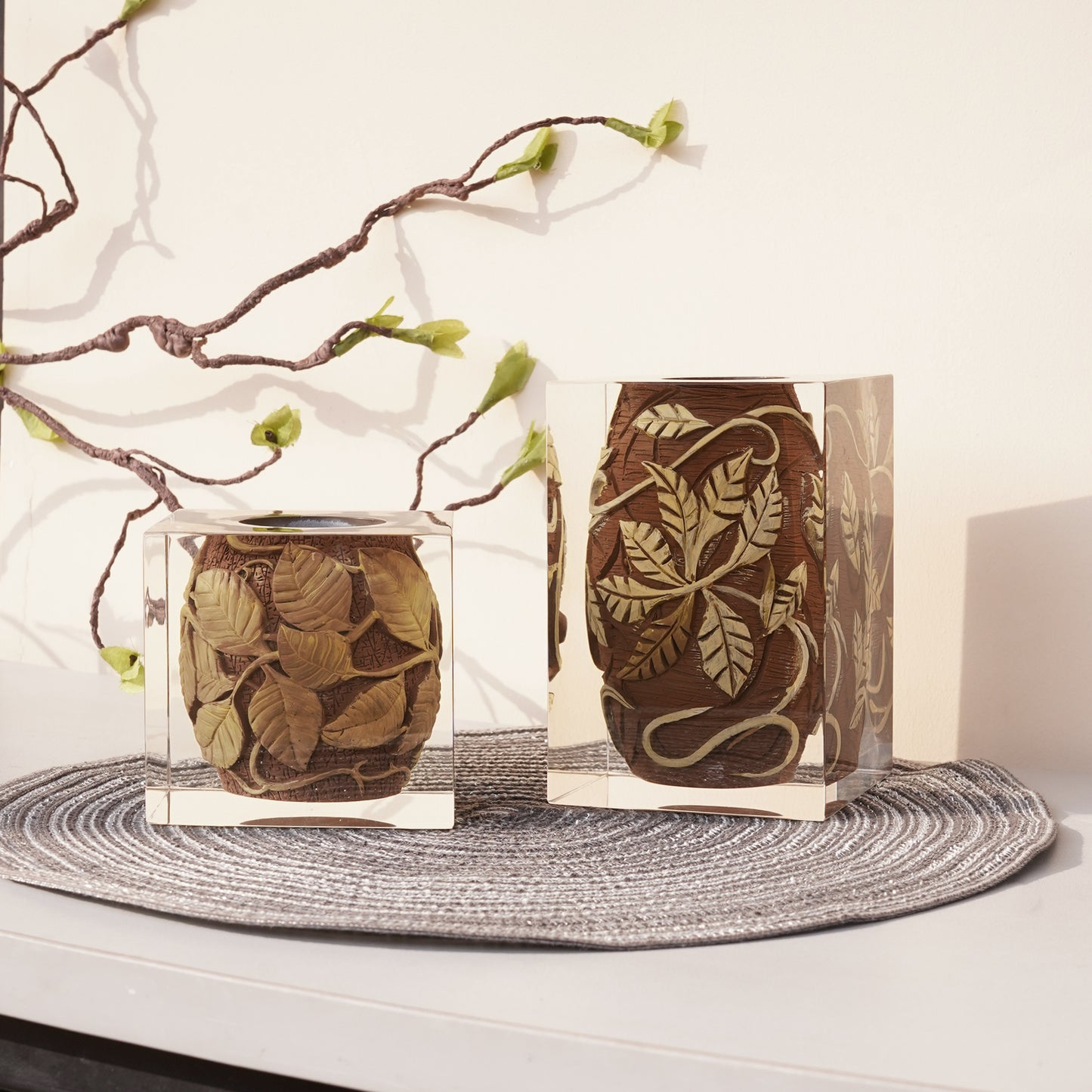 Ceramic small vases featuring rustic embossed leaf vine textures on brown base, encased in clear resin. Tall and short unique creatives vases sitting side by side on a grey mat in a minimalist design interior.