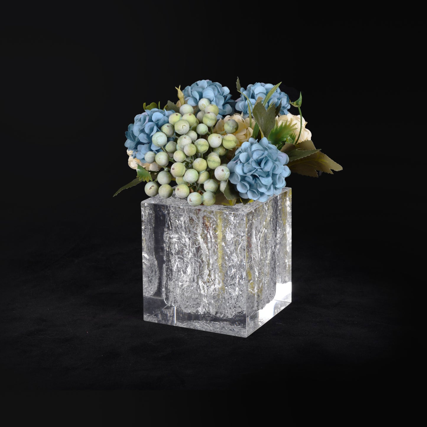 Mini cube vase with ice-like texture filled with blue flowers and berries against dark background. Contemporary artistic vase with a glass-clear body.