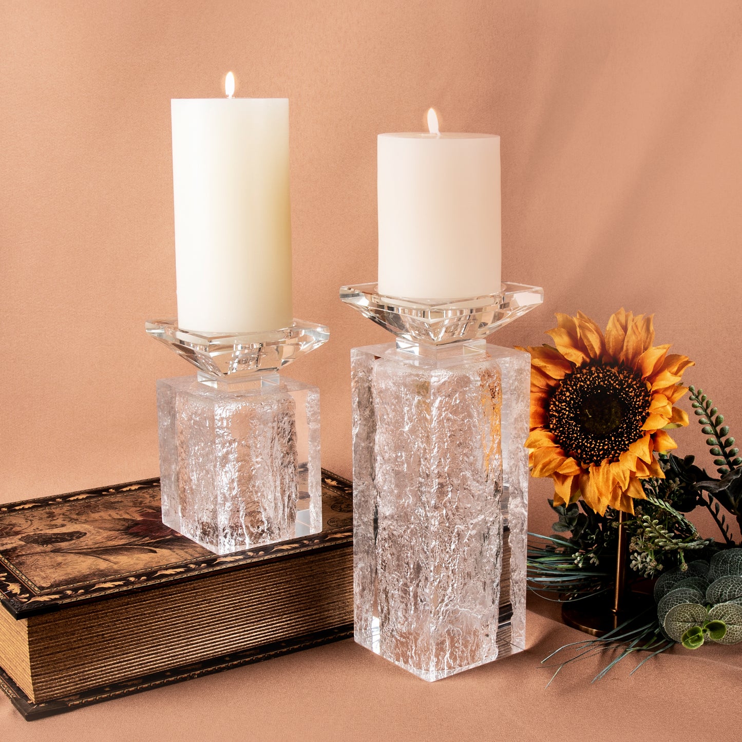 Two decorative candle holders displayed together with lit pillar candles beside decorative sunflower and vintage book. Unique clear acrylic candle holders used as nordic decors for aesthetic room on a orange backdrop.