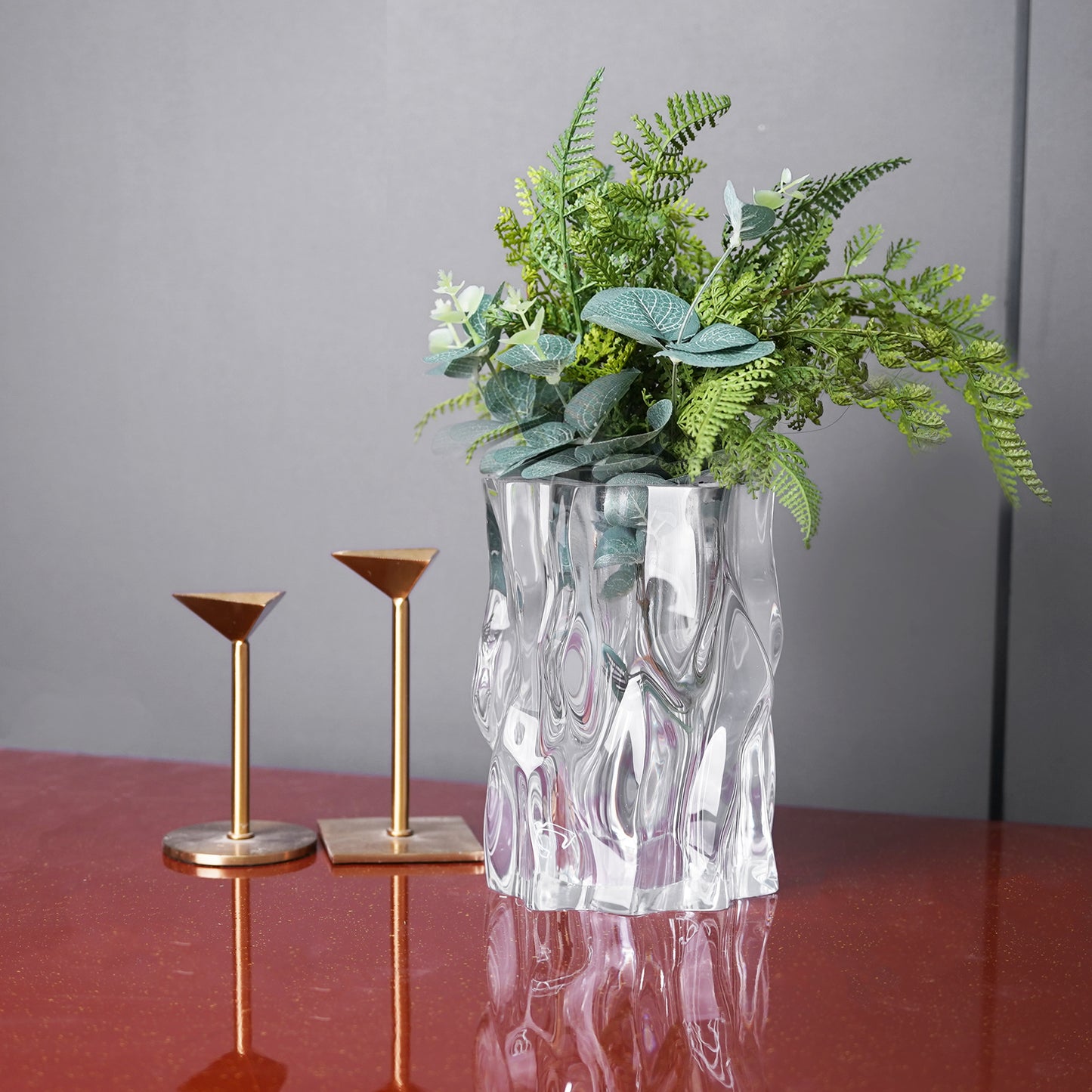 Ripple-textured clear acrylic vase styled with green foliage on a red tabletop. Aesthetic room decor styled with botanics and gold candleholders, placing on red tabletop.